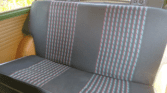 A vintage mini car’s backseat with dark upholstery and vertical striped fabric panels in red, green, and gray. The tan side panels and two speakers on the rear deck stand out as sunlight enters from the left, illuminating part of the seat.