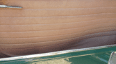 Close-up of a classic mini car door interior with tan, horizontally stitched leather upholstery, a chrome door handle at the top left, and a glossy green metal frame at the bottom, partially reflecting sunlight and gravel on the ground.
