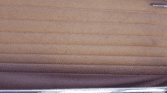 Close-up of a vintage mini car door interior featuring tan padded, horizontally-stitched upholstery. The bottom edge has a shiny chrome trim with green painted metal beneath it. Faint texture and subtle creases are visible on the upholstery.