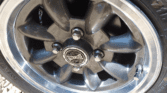 Close-up of a shiny, dark gray alloy wheel with five spokes and visible lug nuts, attached to a mini car tire. The “ULTRALITE” center cap gleams in the sunlight, highlighting the metallic finish against a gravel background.