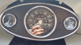 Close-up of a vintage Mini car dashboard with three round gauges. The center speedometer shows up to 100 mph; odometer reads 88154. Left gauge displays temperature, right shows oil and fuel. A hand holding a phone is reflected in the glass.