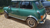 A classic green Mini car with a white roof is parked on a gravel driveway beside a modern gray SUV. The sun shines, reflecting on the chrome details and casting shadows below. Bushes and blue sky provide a scenic backdrop.