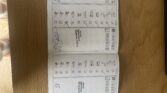 An open mini car service booklet rests on a wooden table, revealing maintenance checklists in German with ticked boxes and signatures. Both pages mirror each other as records—part of a mini trader’s feet appears at the edge.