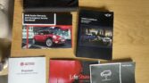 Six MINI car booklets, including owner's manuals, warranty and service handbooks, and a LifeShine booklet, are spread out on a wooden table. A black pouch sits at the top, with part of a cushioned chair visible nearby.
