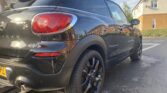 A shiny black Mini Paceman car is parked on a residential street. Captured from the rear left side, this sleek mini car shows off its red taillight, black alloy wheels, and dual exhausts against houses and a clear blue sky.