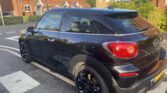 A black MINI Paceman mini car is parked on a residential street near a crosswalk, with red brick houses and greenery in the background. The car features tinted windows, black alloy wheels, and UK plates under sunny, clear skies.