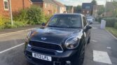 A black Mini Countryman, a classic mini car with the license plate "BT13 LFL," is parked partially on a curb in a suburban neighborhood with brick houses, greenery, and a clear sky. Another car is parked further down the street.