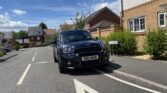 A black Mini car with license plate SR03 ROM is parked partly on a curb and cycle lane beside a residential street on a sunny day. Modern houses and green bushes line the road, beneath a mostly blue sky with white clouds.