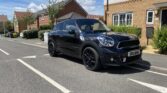 A black Mini Countryman with tinted windows—a stylish mini car—is parked on a suburban street near brick houses and greenery under a blue sky with scattered clouds. The car’s license plate reads “SR13 CRV.” Shadows indicate bright sunlight.