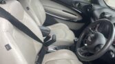 Interior of a MINI car with cream leather seats, black trim, and a manual gear stick. The driver’s seatbelt is fastened. The dashboard displays round air vents and a MINI logo on the steering wheel. Seen through the window: bushes and a silver car—perfect for any MINI trader.