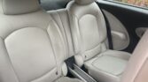 The image shows the back seat area of a mini car with light beige leather upholstery, featuring two bucket-style seats with visible stitching and headrests. Bushes with green and red leaves are visible outside the car window.