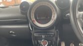 The image shows the interior center console of a MINI car, featuring a circular dashboard display with radio information, climate control buttons, toggle switches, and visible foot pedals below. Houses are visible through the windshield in the background.