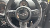 Close-up view of a Mini Cooper steering wheel with the iconic Mini logo in the center. The dashboard displays analog gauges, while control buttons and car pedals add to this classic mini car's sporty interior—perfect for any mini trader.