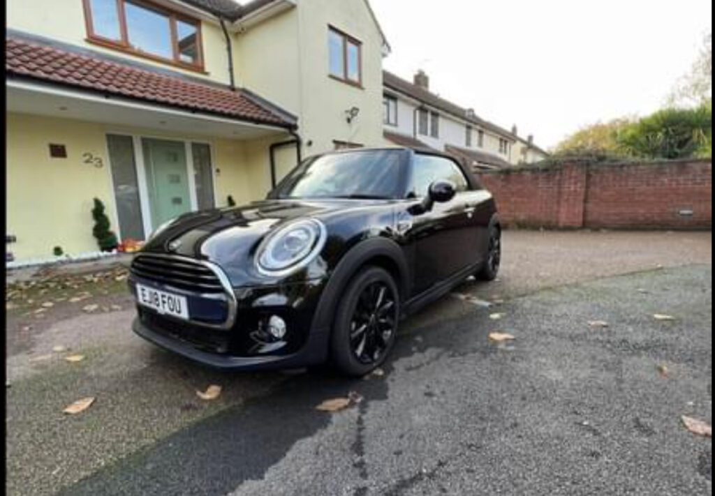 A shiny black mini car convertible with the roof up is parked on a driveway in front of a cream-colored house with brown roof tiles. Fallen leaves are scattered on the ground, and part of a brick wall is visible in the background.