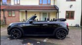 A black convertible mini car with its roof down is parked in front of a cream-colored house with a brown tile roof and several windows. The mini sits on the driveway under clear, dry weather.