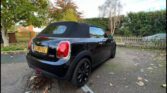 A black mini car convertible with its soft top up is parked on a driveway. The UK license plate reads "EJ18 FOU." Autumn leaves cover the ground, with trees, a wooden fence, and a hedge in the background.