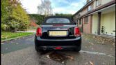 A sleek black Mini convertible—perfect for any mini car enthusiast—is parked on a driveway before a brick house. Fallen leaves dot the ground, the sky is partly cloudy, and the license plate reads "EJ18 PGU.