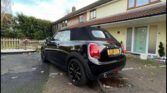 A sleek black Mini convertible with a UK license plate is parked in a driveway beside a modern house with a white door. Wet autumn leaves cover the ground, while trees and a wooden fence frame this stylish mini car scene.