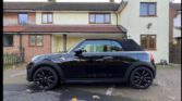 A black Mini Cooper convertible with the roof up is parked on a driveway in front of a cream-colored two-story house. This stylish mini car features black alloy wheels, and a few leaves are scattered on the ground nearby.