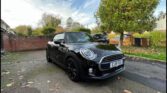 A shiny black Mini convertible with a closed roof is parked on a leaf-strewn driveway. Autumn trees and a brick wall form the backdrop. This stylish mini car, bearing UK plate “EJ18 FDU,” sits under a partly cloudy sky.