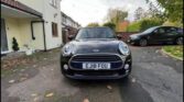 A black mini car with the license plate "EJ18 FOU" is parked on a residential driveway beside a cream-colored house. Another black car is parked further back. The ground has scattered leaves and the sky is mostly cloudy.