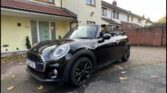 A shiny black Mini Cooper convertible with the top down is parked on a driveway in front of a cream-colored house with a brown tiled roof. This stylish mini car has black wheels, chrome accents, and fallen leaves scattered nearby.