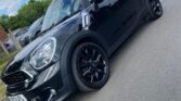 A shiny black Mini Cooper with tinted windows and black rims is parked on a road near other cars. The sky is partly cloudy, and trees and buildings appear in the background, highlighting this stylish mini car in the foreground.