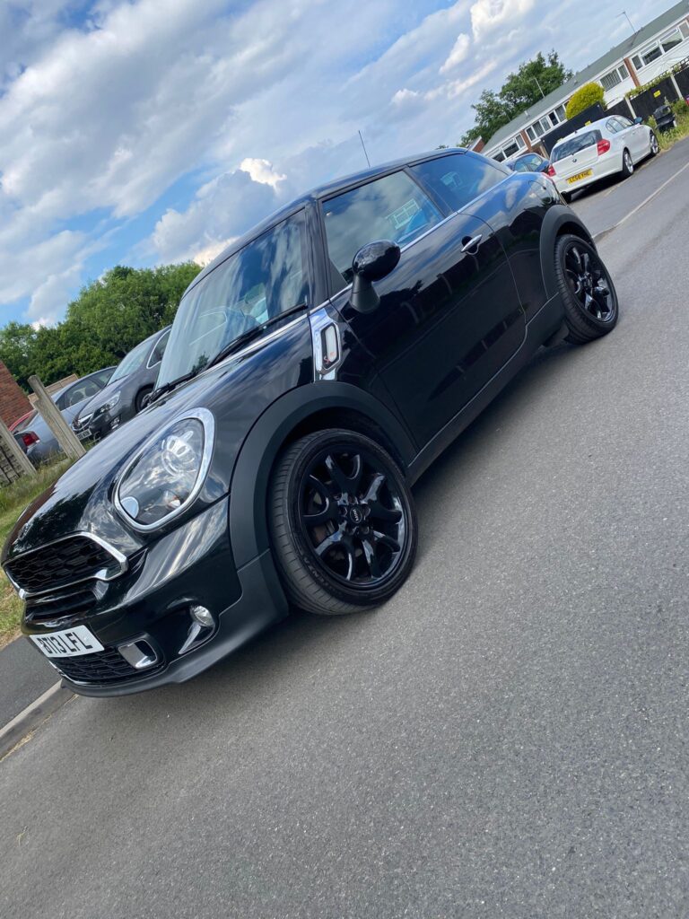A shiny black Mini Cooper with tinted windows and black rims is parked on a road near other cars. The sky is partly cloudy, and trees and buildings appear in the background, highlighting this stylish mini car in the foreground.