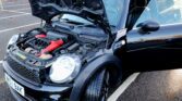 A black Mini car with its hood open reveals the engine and red components. The driver’s door is ajar, and the car is parked in an outdoor lot with wet ground—perfect for any Mini trader seeking a stylish ride.