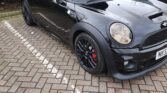 A black Mini Cooper with black alloy wheels and red brake calipers is parked in a lot. The mini car has a roof box attached, foggy headlights, and is angled toward the camera. Part of another black car appears on the right.