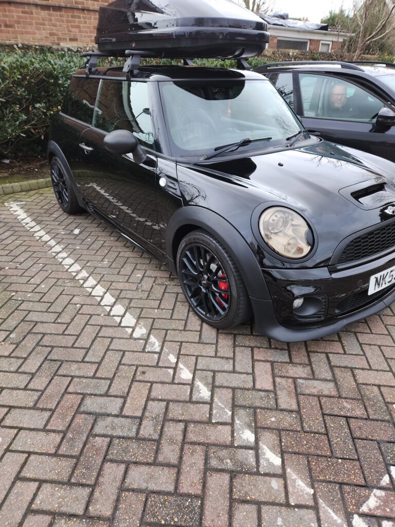 A black Mini Cooper with black alloy wheels and red brake calipers is parked in a lot. The mini car has a roof box attached, foggy headlights, and is angled toward the camera. Part of another black car appears on the right.