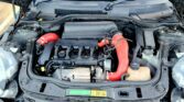 The image shows the engine bay of a Mini with the bonnet open. Visible is a red aftermarket intake pipe, various engine components, and a UK license plate reading "NK59 BAK" at the front of the black vehicle.