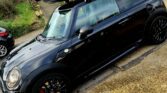 A black mini car with red brake calipers is parked on a residential street, sporting a large black cargo box on its roof. Brick houses and other parked cars line the background under a cloudy sky—a perfect sight for any mini trader.