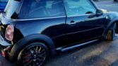 A shiny black Mini car with tinted windows, black alloy wheels, and red brake calipers is parked in a lot. The area features leafless trees, fallen leaves, other cars, and suburban houses in the background. Perfect for a Mini trader!.