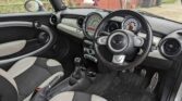 A photo of a right-hand drive mini car interior. It shows a black steering wheel with the Mini logo, large central speedometer, manual gear stick, round air vents, black and light beige seats and pedals, plus a British flag air freshener hanging.