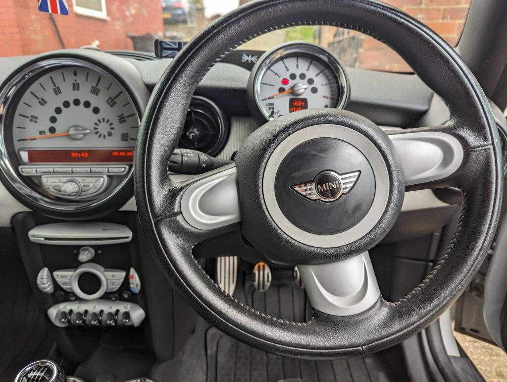 Close-up of a MINI car's interior, highlighting the black steering wheel with the MINI logo. Visible behind it are two round gauges—a large speedometer and a smaller tachometer—along with dashboard buttons and pedals showcased for a mini trader.