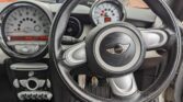 Close-up of a MINI car's interior, highlighting the black steering wheel with the MINI logo. Visible behind it are two round gauges—a large speedometer and a smaller tachometer—along with dashboard buttons and pedals showcased for a mini trader.