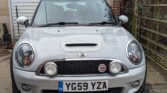 A silver Mini Cooper S mini car is parked in front of a white garage door. The number plate reads "YG59 YZA." The Mini Cooper logo and red "S" badge are on the grille, with greenery and a metal fence to the right.