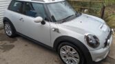 A silver Mini Cooper mini car with a black roof and white racing stripes is parked on a wet concrete driveway next to a metal fence and some bushes. The clean, shiny body reflects the surroundings, making this mini a standout choice for any mini trader.