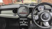 Interior view of a right-hand drive MINI Cooper mini car. The central speedometer and controls are prominent, with a British flag air freshener hanging. The dashboard is black and gray, and the MINI logo is visible on the steering wheel. Brick houses are seen outside.