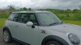 A silver mini car with black accents is parked on a gravel surface. Green grass and trees fill the background under a cloudy, overcast sky. The car is seen from the front left side, showing the front wheel and part of the windshield.