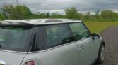 A silver mini car with a black roof is parked on a gravel area beside a grassy field and green trees under an overcast sky. The mini faces away from the camera, displaying its rear and right side.