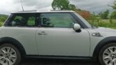 A white mini car with a black roof and side mirrors is parked on a gravel surface near green grass. There are cloudy skies overhead and trees in the background, suggesting a rural setting. The mini is viewed from the side.