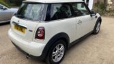 A white Mini Cooper One car, listed on Mini Trader, is viewed from the rear left side and parked on a gravel driveway beside a house with brick and stone walls. The mini car features a black roof, silver alloy wheels, and plate YK61 XNL.