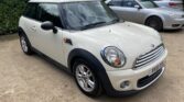 A white Mini Cooper mini car with a black roof and side mirrors is parked on a gravel surface, facing left. The car has chrome accents, clear headlights, and compact, sporty styling. Another silver car and green hedges are visible in the background.