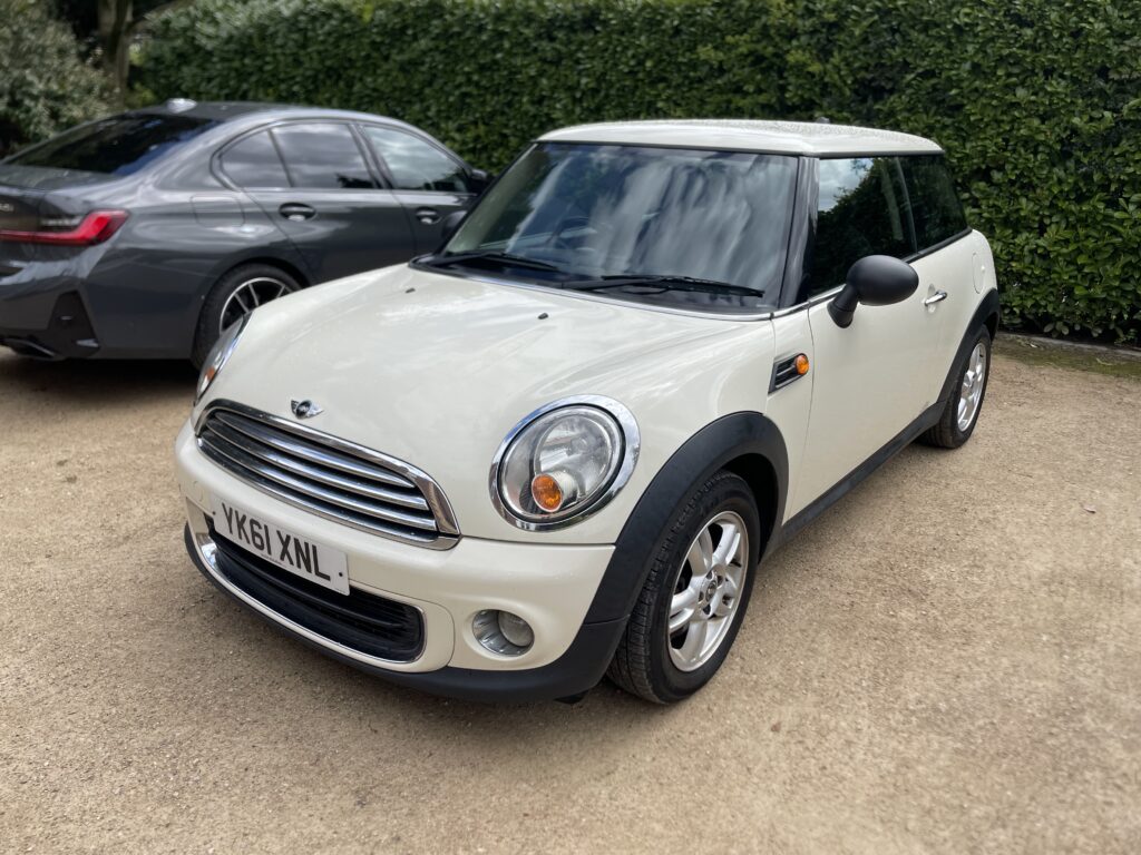 A white mini car with chrome accents is parked on a gravel surface beside a dark grey sedan. Green hedges are in the background, and the UK license plate reads "YK61 XNL," hinting at an outdoor parking area perfect for any mini trader.