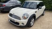 A white mini car with chrome accents is parked on a gravel surface beside a dark grey sedan. Green hedges are in the background, and the UK license plate reads "YK61 XNL," hinting at an outdoor parking area perfect for any mini trader.