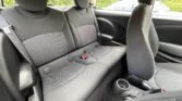 The image shows the back seat of a mini car with black fabric upholstery, three seatbelt buckles, and a center armrest. The front passenger seat is partially visible. Outside the window, greenery and a white car can be seen.