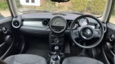 Interior view of a Mini car, showing two front seats, a rounded dashboard with a large central speedometer, a steering wheel with the Mini logo on the right, and circular air vents. The outside garden and part of a brick house are visible through the windshield.