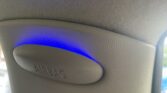 Close-up of a mini car’s interior pillar with an "AIRBAG" cover on textured grey material. A blue light above casts a curved glow on the trim, while part of a window reveals a blurry view outside—perfect for any mini trader’s keen eye.